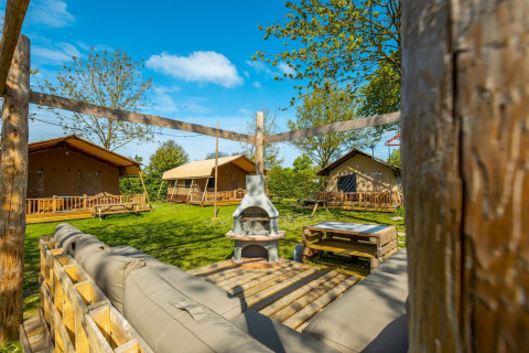 Vue extérieure d’un lodge avec des tentes, bancs en bois, pelouse verte et coin salon sous le ciel bleu.