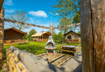 Vista esterna di un lodge con tende, panche in legno, prato verde e zona relax sotto un cielo sereno.