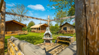 Vue extérieure d’un lodge avec des tentes, bancs en bois, pelouse verte et coin salon sous le ciel bleu.