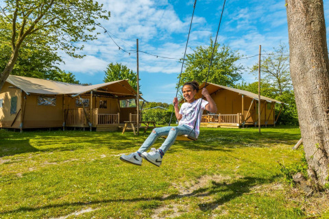 A child enjoys swinging in front of safari tents at a lodge, surrounded by greenery on a bright sunny day.