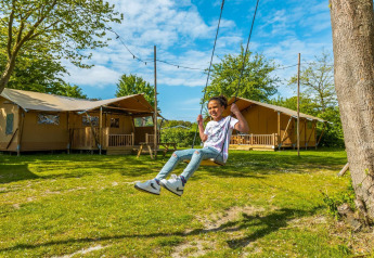 Un enfant se balance devant des tentes safari à un lodge, entouré de verdure sous un ciel bleu et ensoleillé.