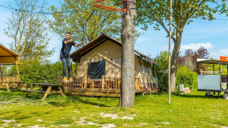 Hombre monta en tirolesa frente a una cabaña en un día soleado, rodeado de árboles y césped verde.