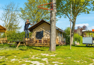 Mann schwingt sich an einer Seilrutsche vor einer Hütte, umgeben von Bäumen und grünem Gras im Sonnenschein.