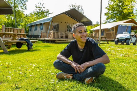 Jeune homme assis en tailleur, souriant sur l’herbe devant des tentes et une voiturette à un lodge.
