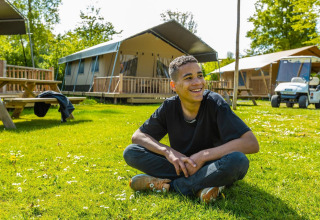 Young man sits cross-legged and smiling on the grass near tents and a golf cart at a sunny lodge.