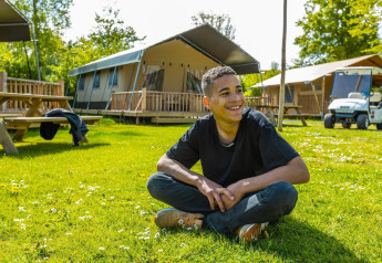 Jonge man zit glimlachend op het gras bij tenten en een golfkar op een zonnige dag bij een lodge.