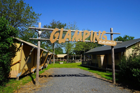 Entrance to a glamping lodge, featuring large tents, greenery, and a wooden sign reading 'GLAMPING'.