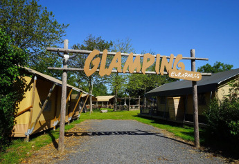 Ingresso di un glamping lodge con tende lussuose, erba verde, alberi e il grande cartello 'GLAMPING'.