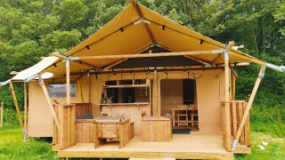 Glampingtent Dreamer con porche de madera, zona de asientos al aire libre y rodeado de árboles verdes.