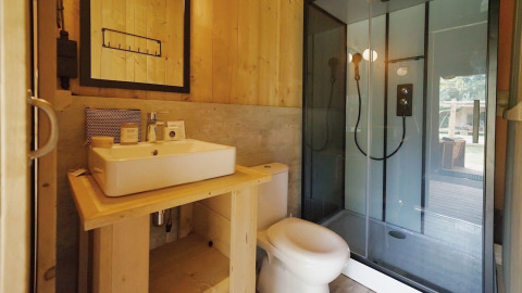 Moderne badeværelse med trævægge, hvid håndvask, toilet og glasbrusekabine i Glampingtent Sunshine.