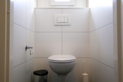 Toilettes modernes d'un lodge avec WC suspendu, poubelle, brosse et murs carrelés blancs.