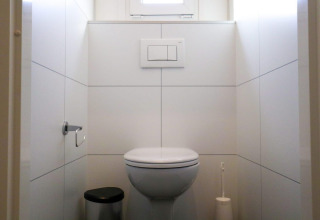 Bagno moderno di lodge con WC sospeso al muro, pattumiera, scopino e pareti piastrellate bianche.