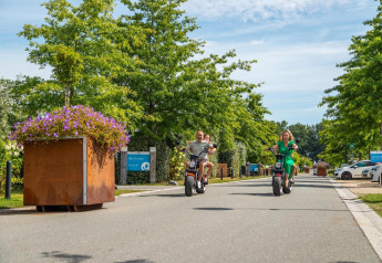 Twee volwassenen en twee kinderen rijden op elektrische scooters door een groene glampingstraat.