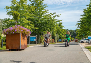 Deux adultes et deux enfants roulent en scooter électrique sur une route arborée d’un site de glamping.