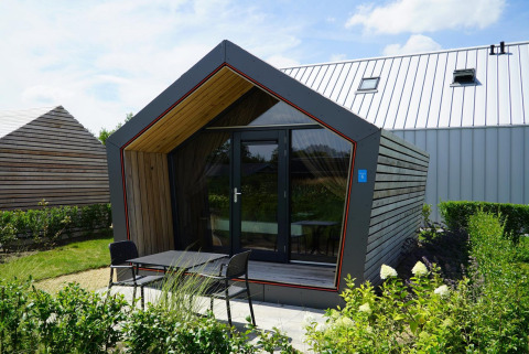 Eine moderne Solo Retreat-Lodge mit Holzfassade, Glasfront und Terrasse im Grünen an einem sonnigen Tag.