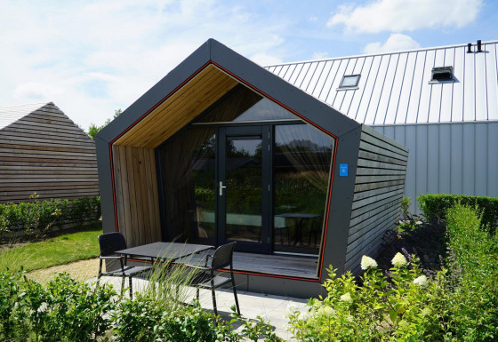 Eine moderne Solo Retreat-Lodge mit Holzfassade, Glasfront und Terrasse im Grünen an einem sonnigen Tag.