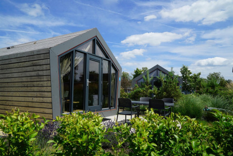 Moderne Solo Retreat-lodge met grote ramen en terrasstoelen, omgeven door groene planten en blauwe lucht.