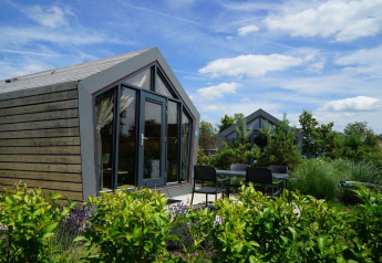 Moderne Solo Retreat-lodge met grote ramen en terrasstoelen, omgeven door groene planten en blauwe lucht.