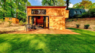 A bright modern tiny house with wooden exterior, patio and lawn, nestled in a lush green forest landscape.