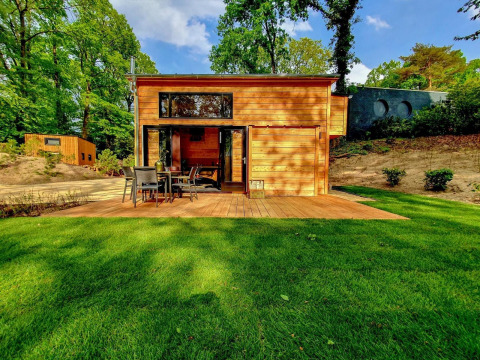 Ein modernes Tiny House aus Holz mit großer Terrasse und Garten, gelegen in einer grünen, bewaldeten Umgebung.