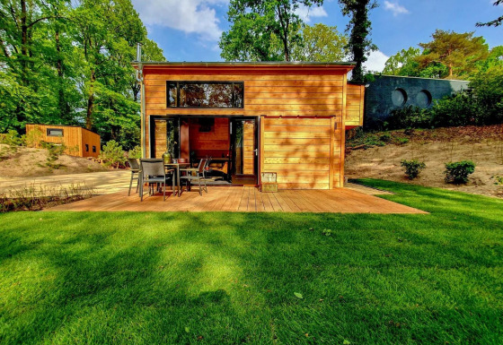 Een modern houten tiny house met terras en gazon, gelegen in een groene, bosrijke omgeving.
