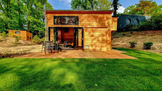 En lyst og moderne tiny house lavet af træ, omgivet af grøn græsplæne og skovområde med udendørs terrasse.