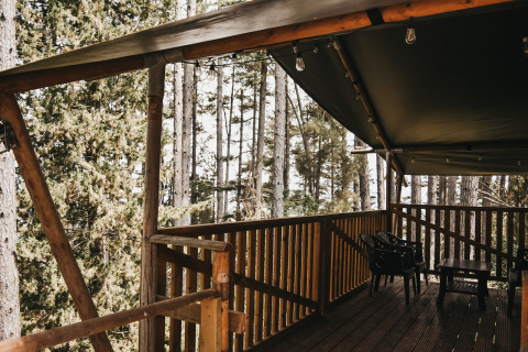 Holzveranda mit schwarzem Zeltdach, Kunststoffstühlen und Tisch, umgeben von italienischem Wald.