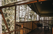 Terraza de madera con techo de lona negra, sillas de plástico y mesa entre altos pinos en Italia.