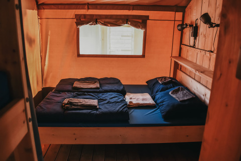 Gemütliche Glamping-Unterkunft mit Holzwänden, Doppelbett, dunkelblauer Bettwäsche und Fenster.