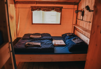 Cómoda habitación de glamping con interior de madera, cama doble, sábanas azul marino y ventana.