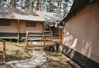 Tienda safari con terraza de madera y sendero en Camping Luna del Monte, rodeada de árboles altos en Italia.