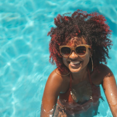 Mujer con cabello rizado y gafas de sol sonriendo en una piscina en un parque vacacional con glamping.