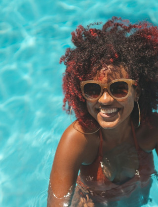 Woman with curly hair and sunglasses smiling in a swimming pool at a holiday park offering glamping.