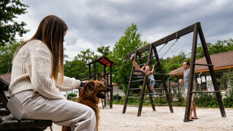 Family at Camping Etxarri holiday park in Navarra, Spain, kids on swings, woman with dog, cloudy sky.