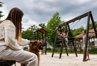 Family at Camping Etxarri holiday park in Navarra, Spain, kids on swings, woman with dog, cloudy sky.