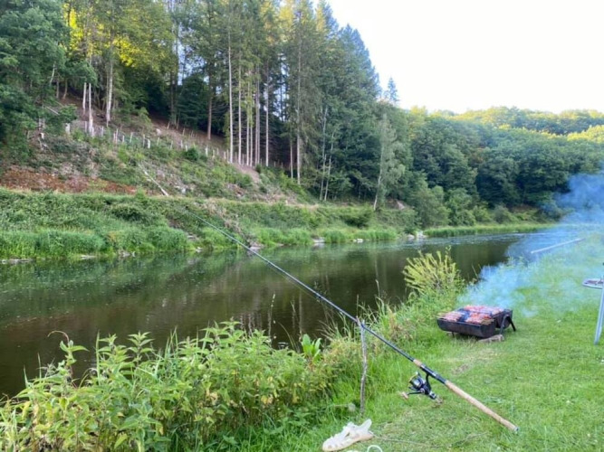 Angelplatz mit Angelrute und Grill am Fluss bei Huttopia Vallée de la Semois, Namur, Belgien, umgeben von Wald.