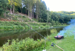 Lugar de pesca con caña y parrilla junto al río en Huttopia Vallée de la Semois, Namur, Bélgica, rodeado de bosque.