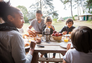 En familie nyder morgenmad udendørs ved et træbord på Huttopia Vallée de la Semois-feriepark i Belgien.