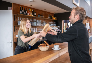 Een lachende medewerkster geeft een vers stokbrood aan een klant aan de toog bij Huttopia Vallée de la Semois.