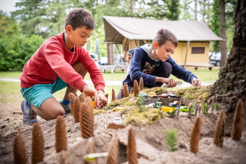 Dos niños juegan al aire libre con coches de juguete y piñas en la arena cerca de tiendas de campaña.