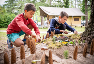 Twee jongens spelen buiten met speelgoedautootjes en dennenappels in zand, bij tenten op een vakantiepark.
