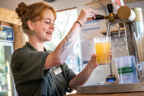 Mujer con placa de nombre sirviendo cerveza de barril y sonriendo en Huttopia Vallée de la Semois, en Bélgica.