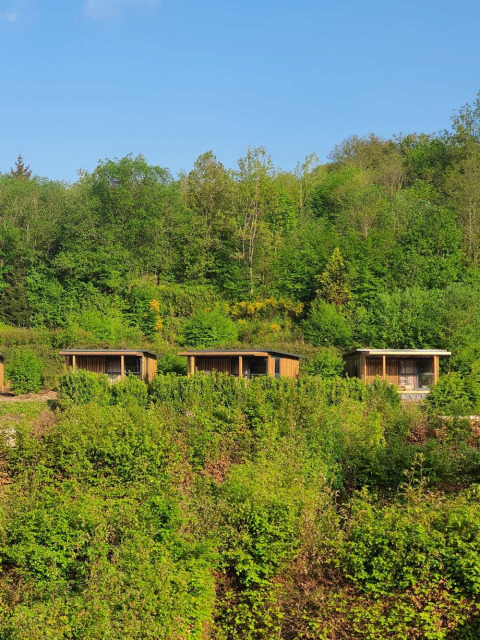 Trois cabanes en bois au cœur de la verdure à Huttopia Vallée de la Semois, Namur, Belgique, sous un ciel bleu.