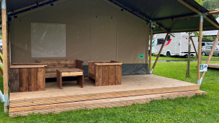 A safari tent with wooden furniture on the porch, surrounded by green grass and RVs in the background.