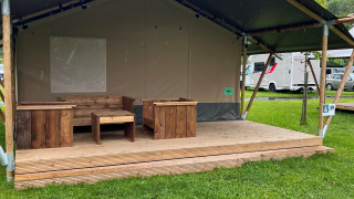 Una tienda tipo safari con muebles de madera en el porche, rodeada de césped verde y caravanas al fondo.