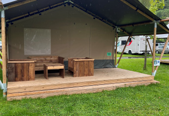 En safaritelt med træmøbler på en veranda, omgivet af grønt græs og campingvogne i baggrunden.