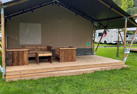 Una tenda safari con mobili in legno sulla veranda, circondata da erba verde e camper sullo sfondo.