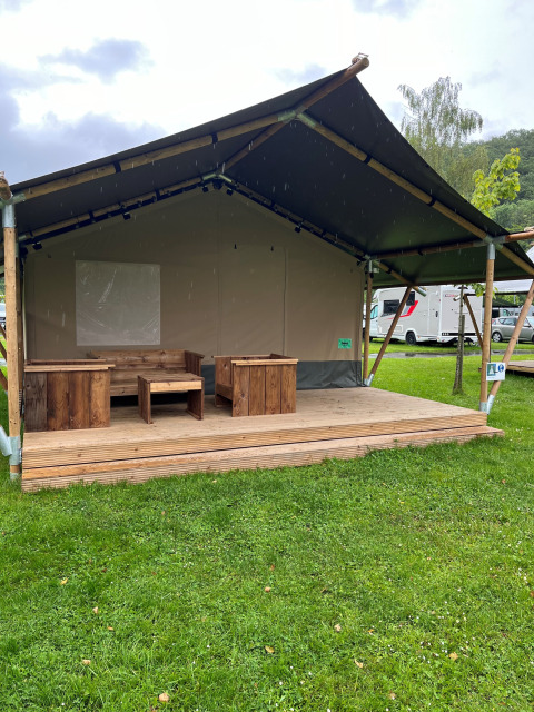 Ein Safarizelt mit Holzmöbeln auf einer Veranda, umgeben von grünem Gras und Wohnmobilen im Hintergrund.