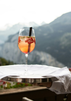 A waiter serves a refreshing cocktail with fruit on a tray, mountains and glamping scenery in the background.