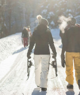 Dos personas caminan por una carretera nevada en un parque vacacional con glamping, al sol de invierno.